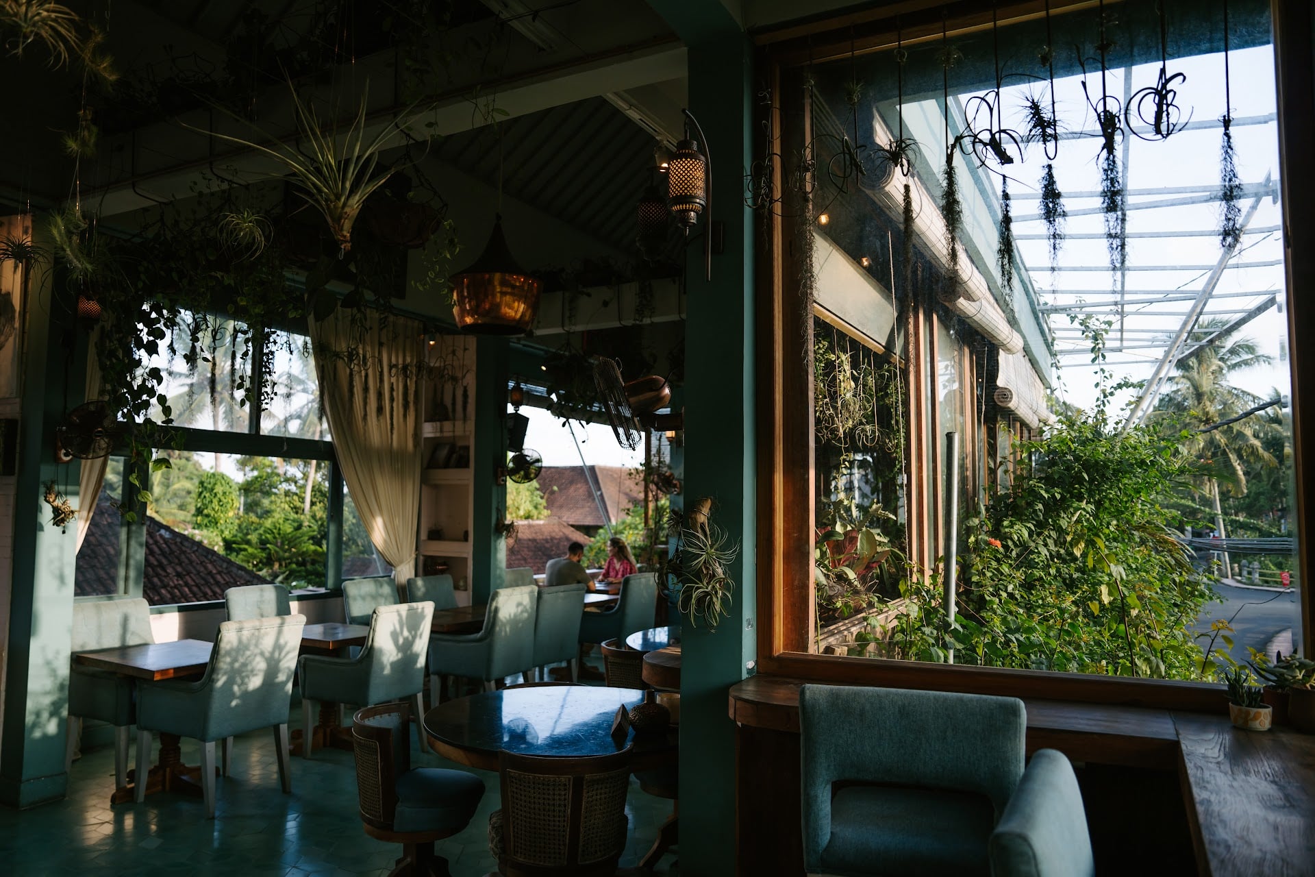 Sunlit dining room at Muse Café & Art with tables by the window overlooking Ubud's tiled rooftops and palm trees