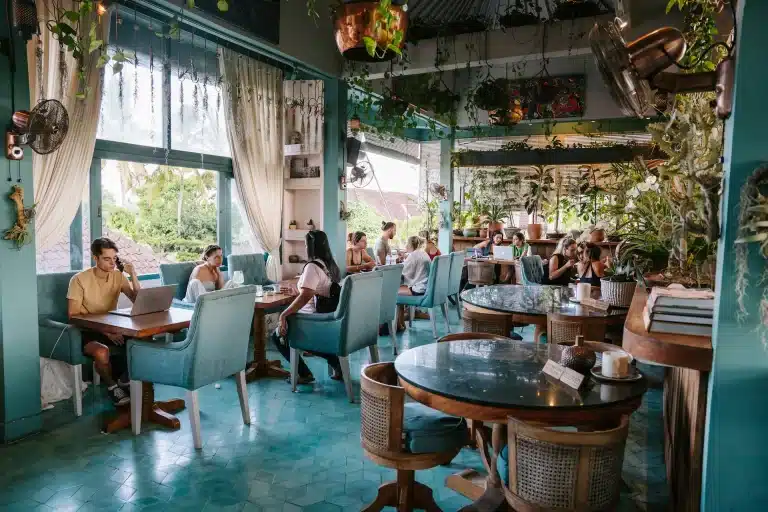 Morning at Muse Café & Art in Ubud: guests at teal armchairs and marble tables among plants, with a window view of trees and rooftops